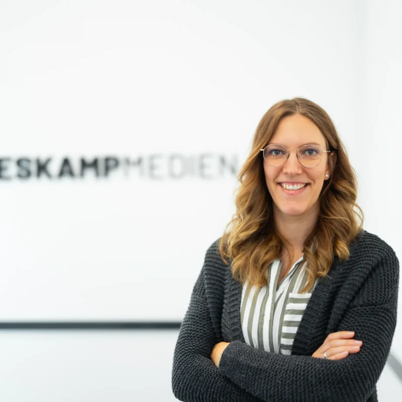 Eine Frau mit gewelltem, hellbraunem Haar und Brille steht lächelnd mit verschränkten Armen vor einer weißen Wand mit dem Logo von Heskamp Medien und dem Text Jaron Heskamp Digitale Visitenkarte.