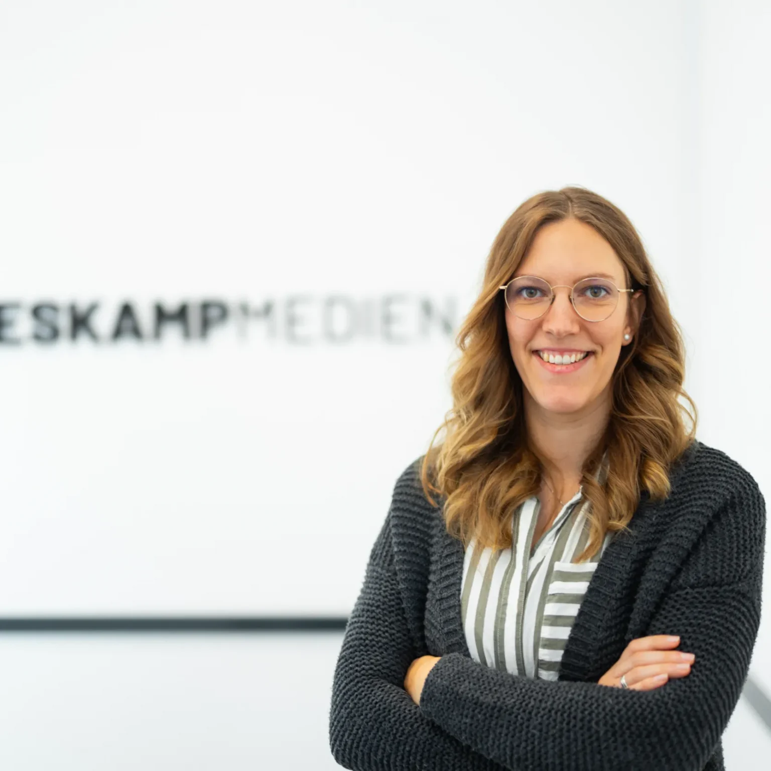 Eine Frau mit gewelltem, hellbraunem Haar und Brille steht lächelnd mit verschränkten Armen vor einer weißen Wand mit dem Logo von Heskamp Medien und dem Text Jaron Heskamp Digitale Visitenkarte.