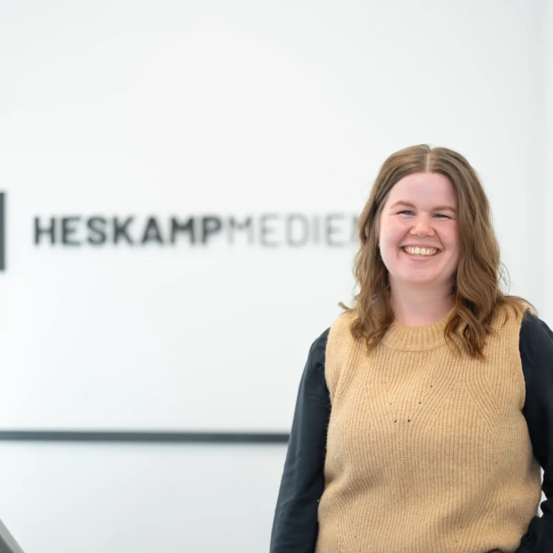 Eine lächelnde Frau, Christin Elfering, mit gewellten braunen Haaren steht in einem Raum neben einer Wand mit dem Logo der Firma Heskamp Medien. Sie trägt einen hellbraunen ärmellosen Pullover über einem schwarzen Hemd, in einem hellen, minimalistischen Raum - ihr Digitale Visitenkarte-Moment.