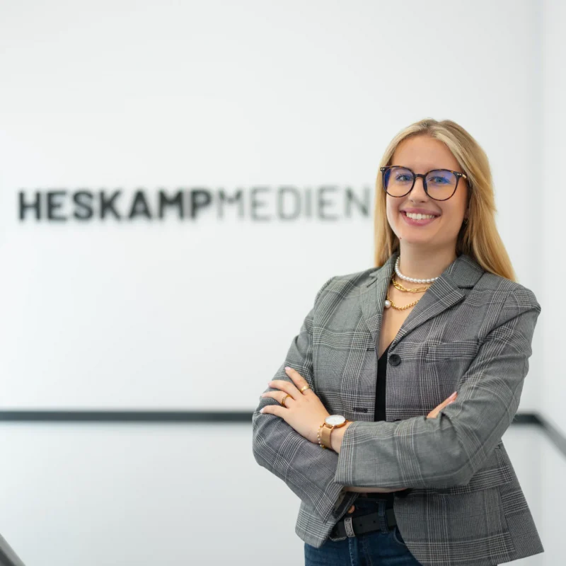 Eine Frau mit Brille, grau-kariertem Blazer und Goldschmuck steht lächelnd mit verschränkten Armen vor einer weißen Wand mit dem Logo "Heskamp Medien".