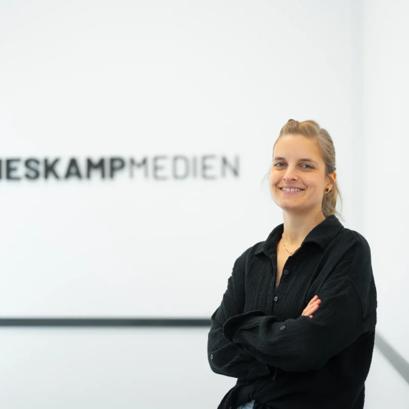 Eine Person mit langen blonden Haaren, die ein schwarzes Hemd trägt, steht lächelnd mit verschränkten Armen vor einer verschwommenen Wand, auf der das Logo von Heskamp Medien und die Carina Ruddeck Digitale Visitenkarte abgebildet sind.