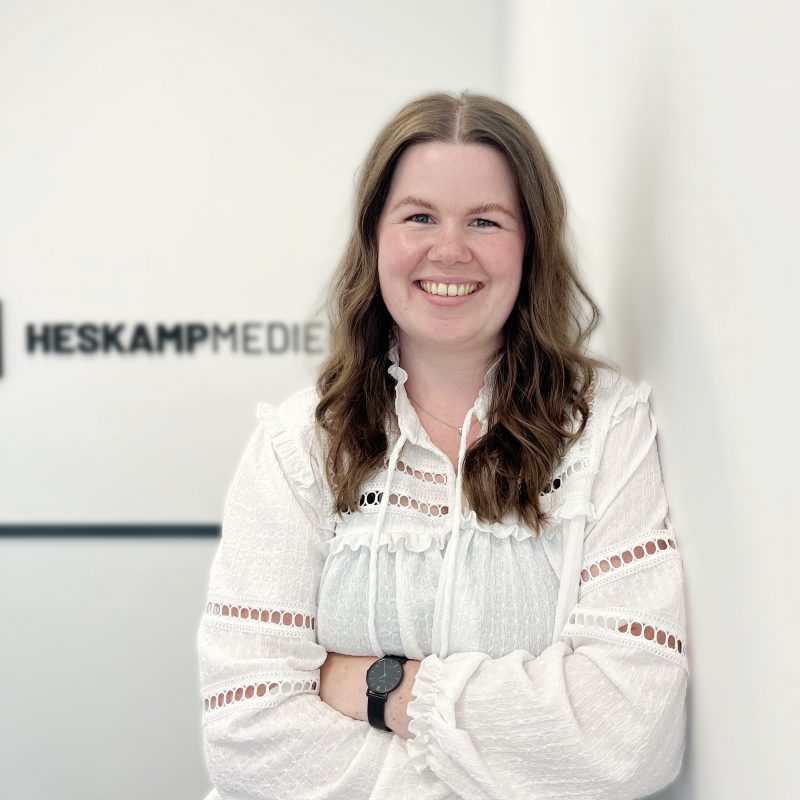 Eine Frau mit langen braunen Haaren, weißer Bluse und schwarzer Uhr steht lächelnd mit verschränkten Armen vor einer weißen Wand mit dem Logo von HESKAMP MEDIA.