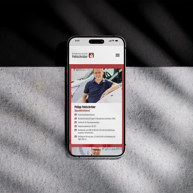 Ein Smartphone, auf dem das Profil der Brandschutztechnik Pohlschröder-Website einer lächelnden Person in einem blauen Hemd neben einem Auto angezeigt wird, mit Text und Symbolen darunter, platziert auf einer strukturierten Oberfläche mit diagonalen Schatten.