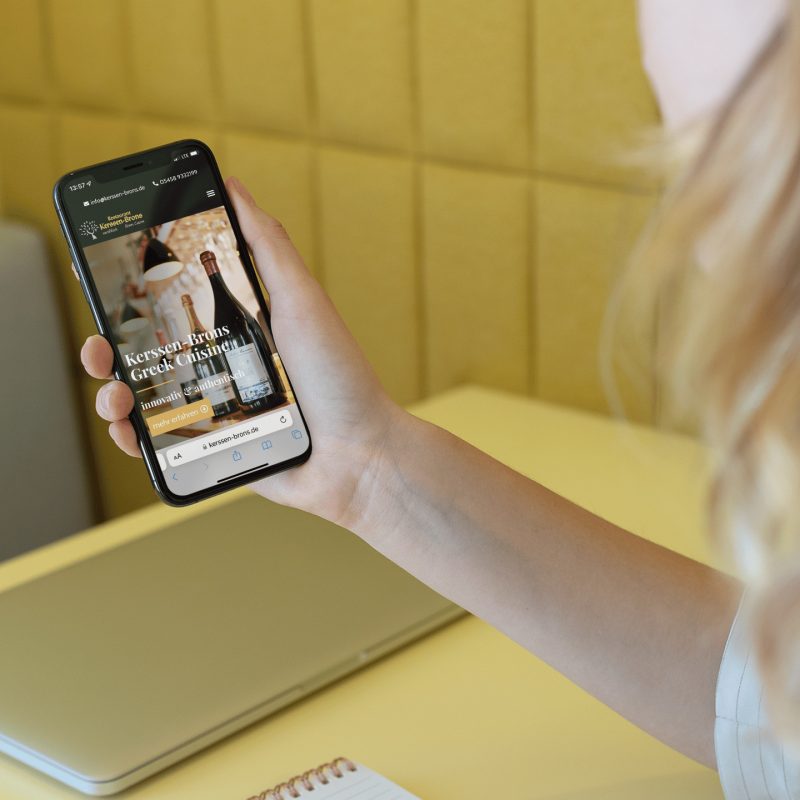 Eine Person hält ein Smartphone in der Hand, auf dem eine Hotelbuchungs-Website angezeigt wird, und sitzt an einem gelben Schreibtisch mit einem geschlossenen Laptop, einem Spiralnotizblock und einem Stift in der Nähe.