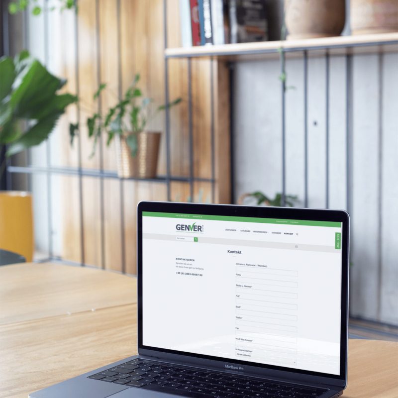 Ein Laptop, auf dem eine Website mit einem Kontaktformular angezeigt wird, steht auf einem runden Holztisch in einem modernen Raum mit Grünpflanzen und Regalen im Hintergrund.